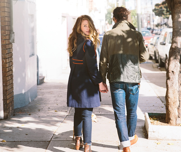 Couple walking along sidewalk holding hands with woman looking behind her